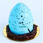 Blue mirror glaze cheesecake shaped like a robin egg with cocoa speckles and piped chocolate nest