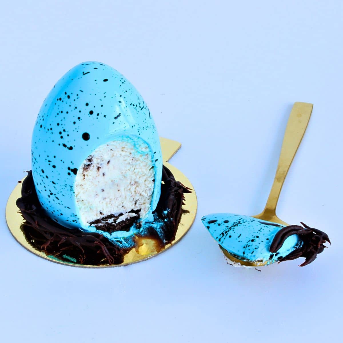 Speckled Robin’s Egg Cheesecake with blue mirror glaze and chocolate ganache nest on gold cake board