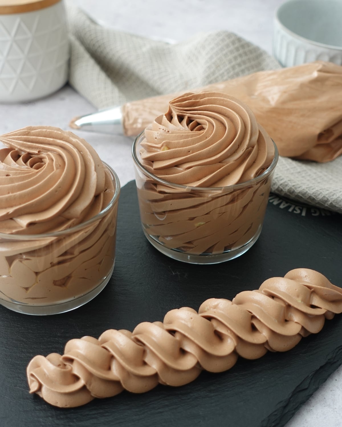 Chocolate hot water buttercream piped into swirls in glass jars with a rope border on a slate board