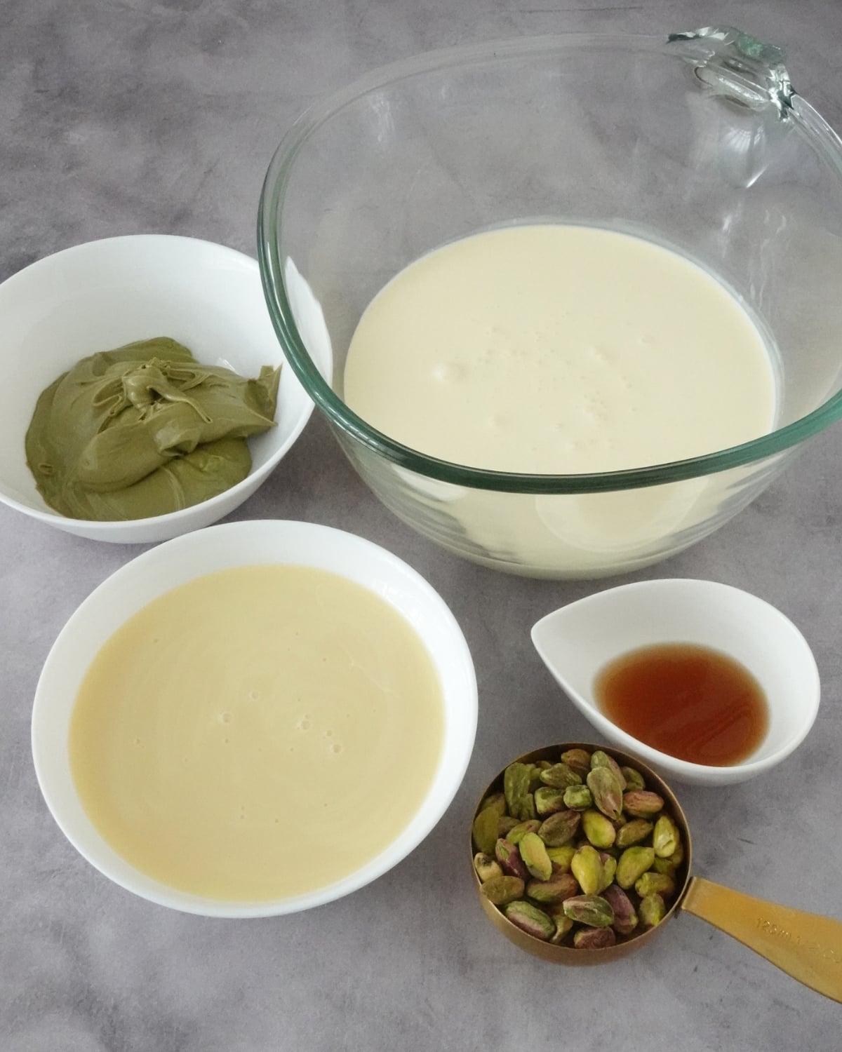 Ingredients for pistachio ice cream including pistachio cream, condensed milk, heavy cream, vanilla, and pistachios in bowls on a grey background