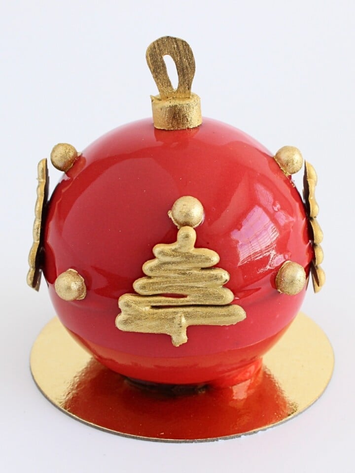 Shiny red mirror glaze Christmas ornament dessert with gold chocolate decorations on a small gold cake board.