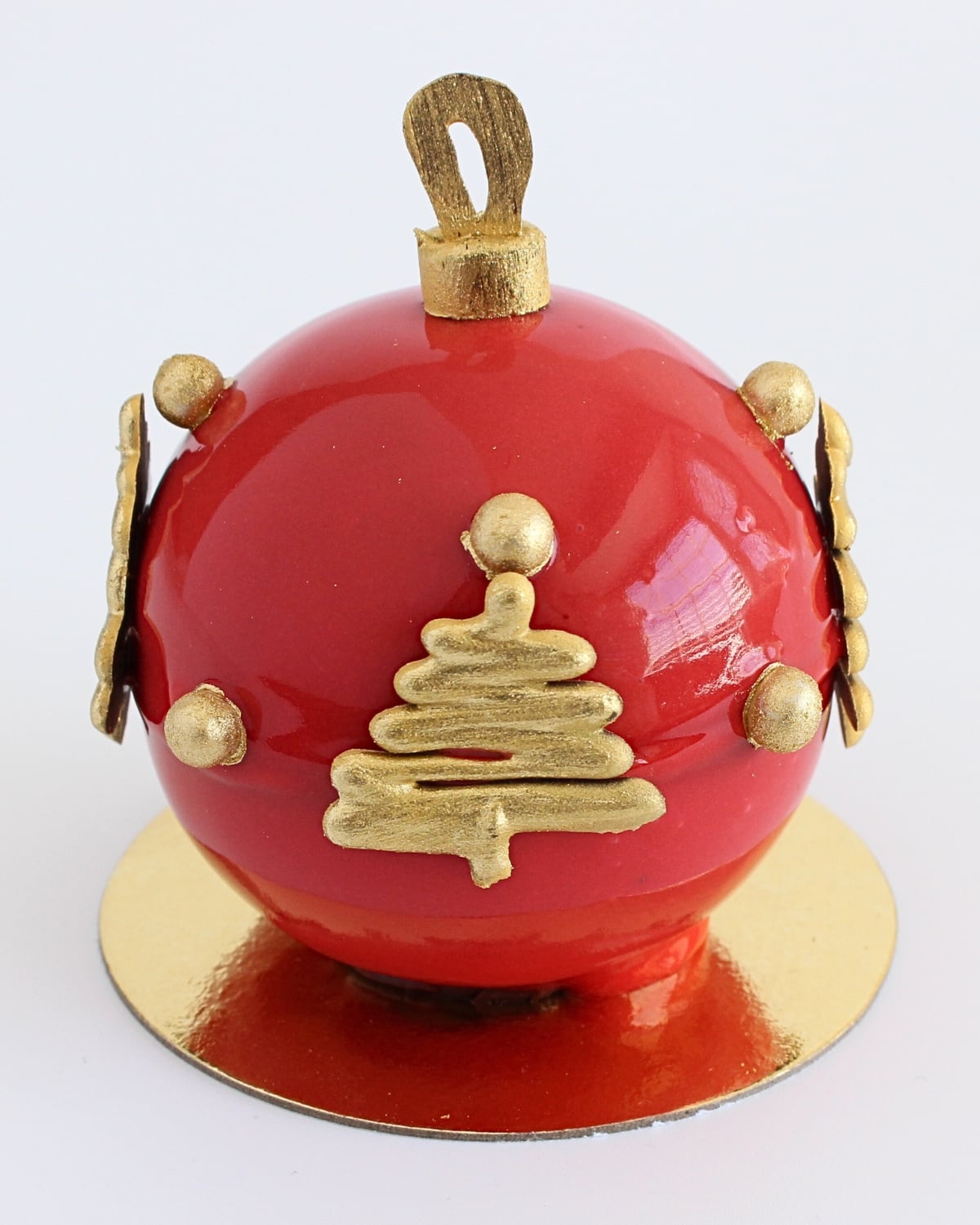 Red mirror glaze Christmas ornament dessert with gold chocolate decorations on a round gold cake board.