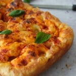 Baked focaccia pizza base on a wooden board topped with melted bocconcini, pizza sauce, and fresh basil leaves