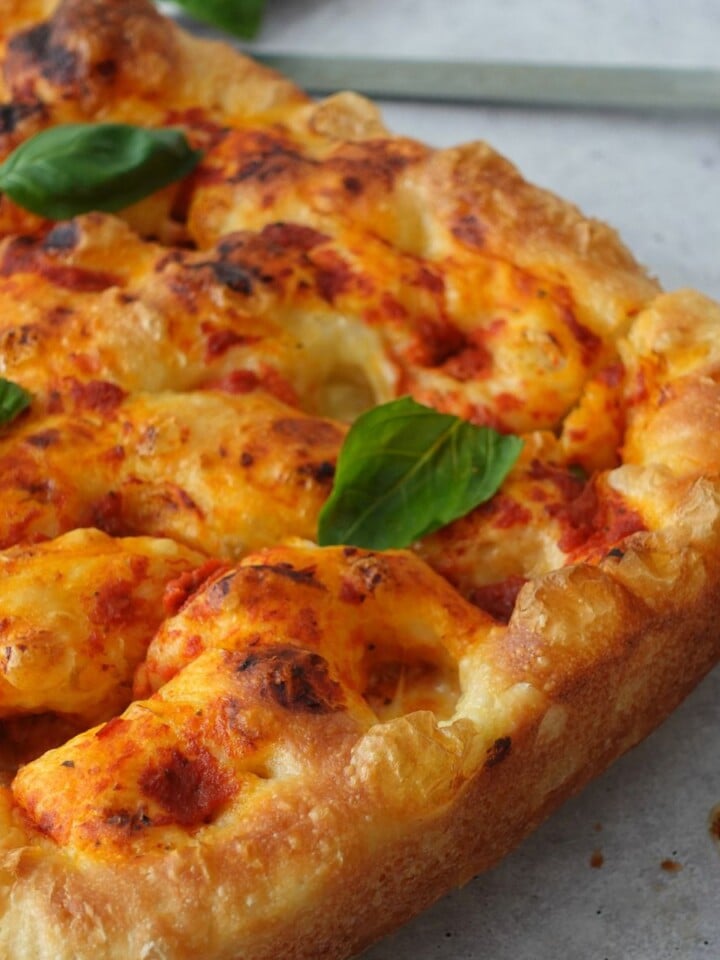 Baked focaccia pizza base on a wooden board topped with melted bocconcini, pizza sauce, and fresh basil leaves