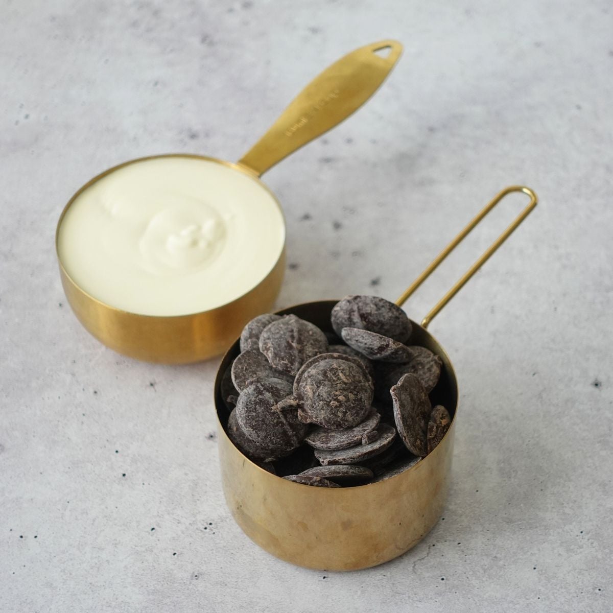 Heavy cream in a gold measuring cup and dark chocolate discs in a gold measuring cup on a grey surface