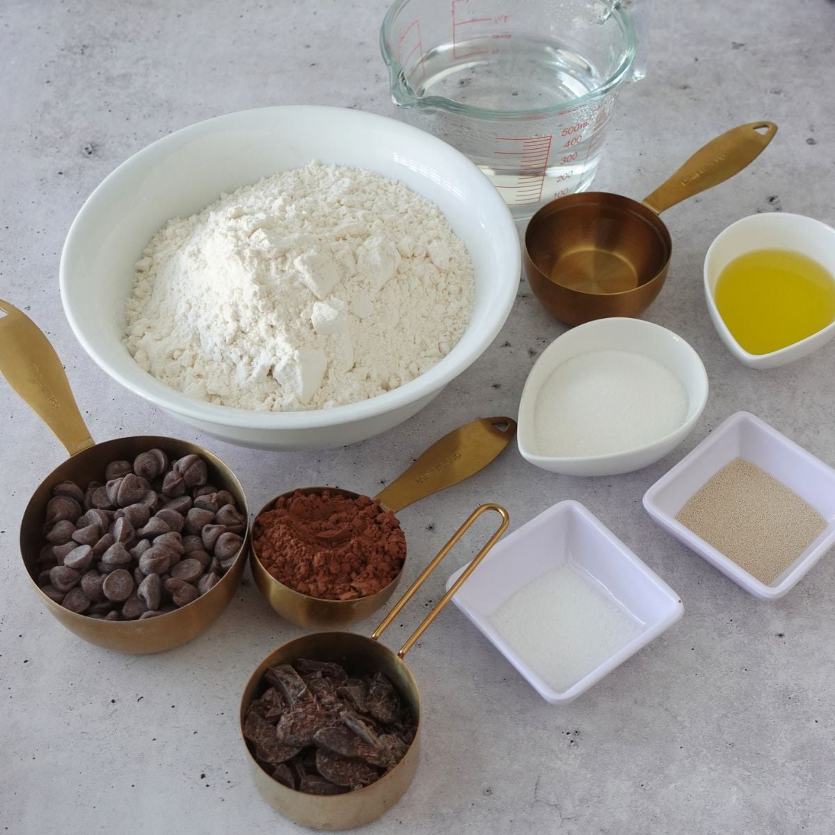 Bread flour, cocoa powder, chocolate chips, chocolate chunks, water, olive oil, sugar, salt, and instant dry yeast laid out on a grey surface