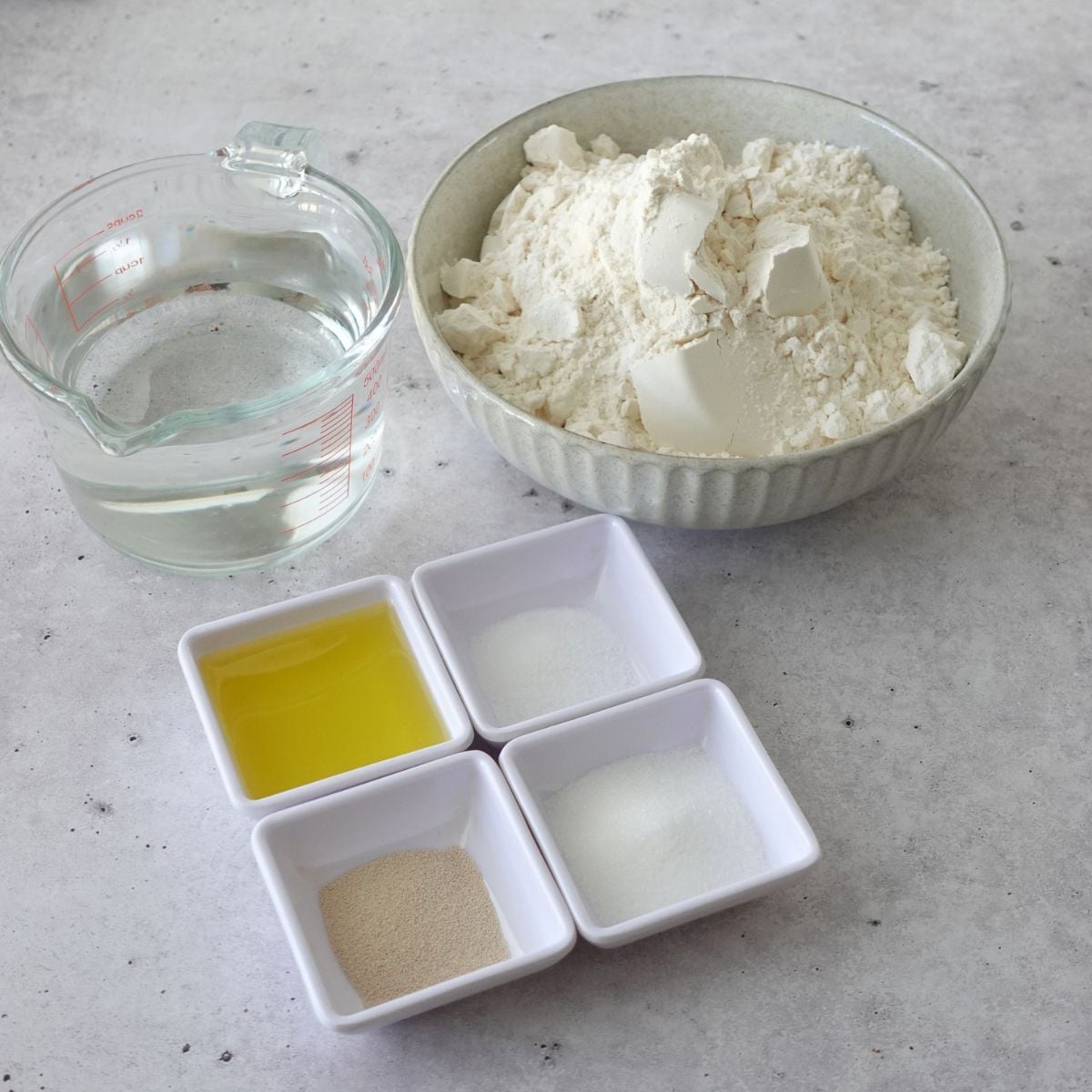 Focaccia pizza base ingredients including bread flour, water, olive oil, instant dry yeast, salt and sugar laid out on a grey surface