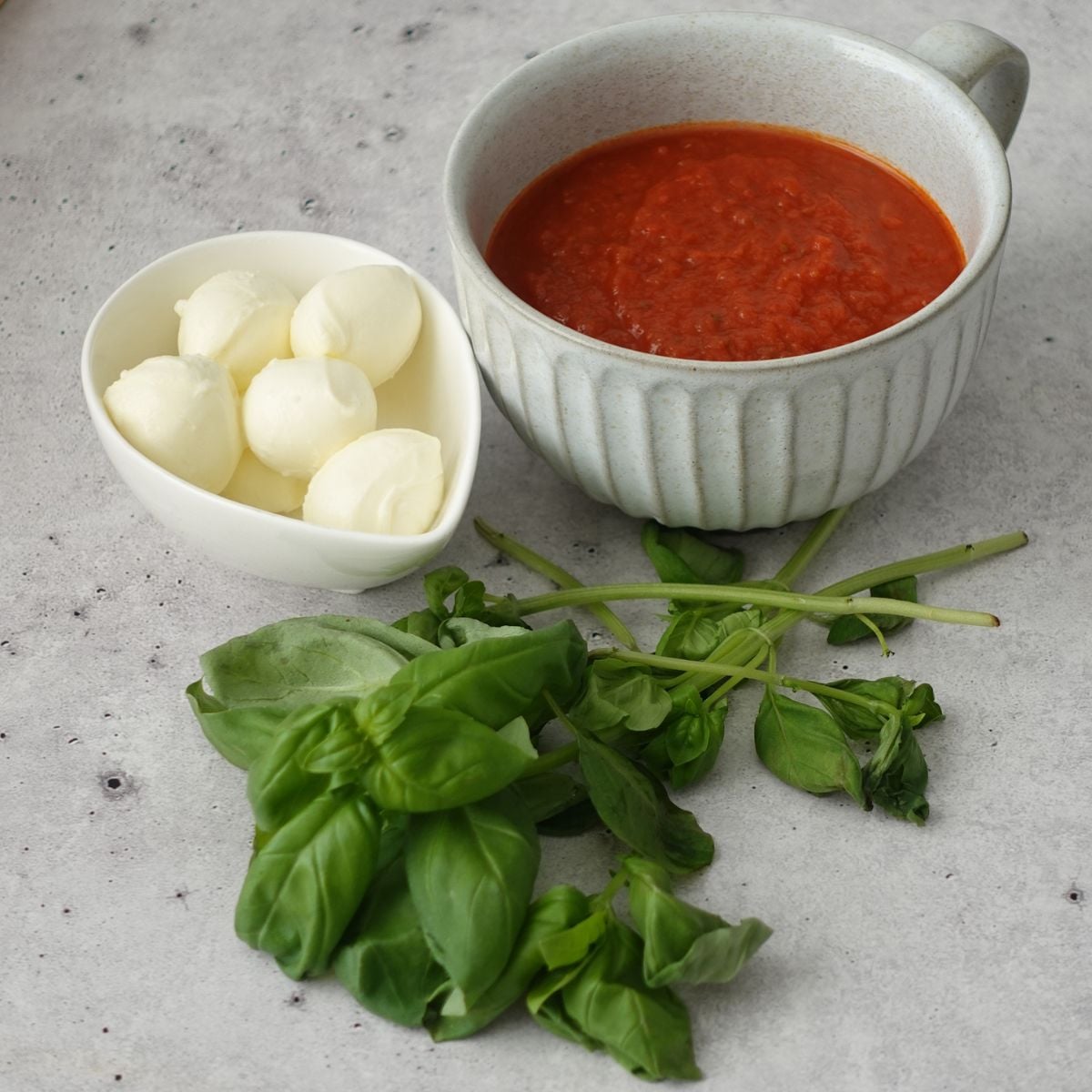 Pizza sauce in a ceramic bowl, mini bocconcini in a white dish, and fresh basil on a grey surface
