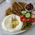 Labneh dip recipe on a platter with za'atar manoosh, cherry tomatoes, cucumber, olives, and cornichons
