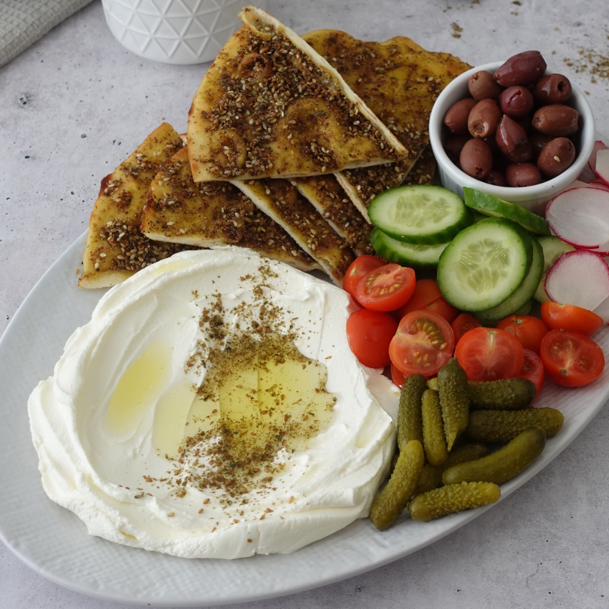 Labneh dip recipe on a platter with za'atar manoosh, cherry tomatoes, cucumber, olives, and cornichons