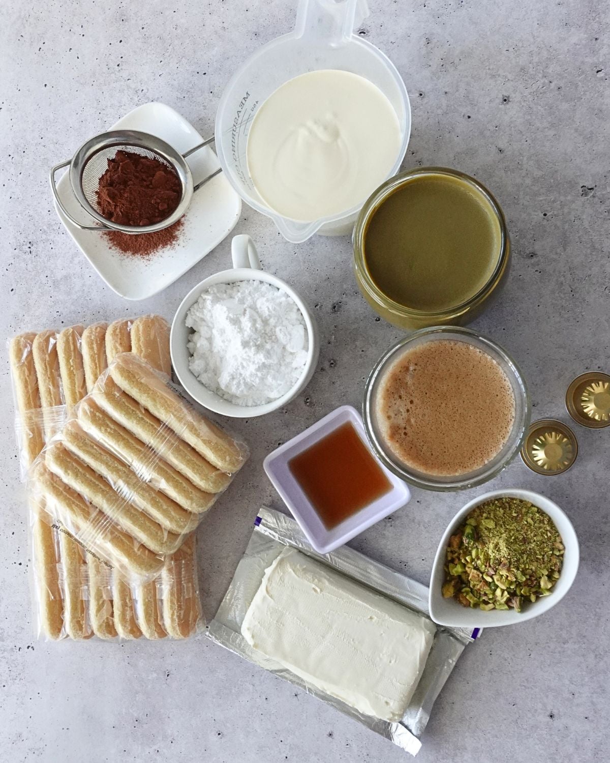 Ingredients for no-bake pistachio tiramisu cheesecake, including ladyfingers, cream cheese, pistachio cream, crushed pistachios, coffee, and cocoa powder