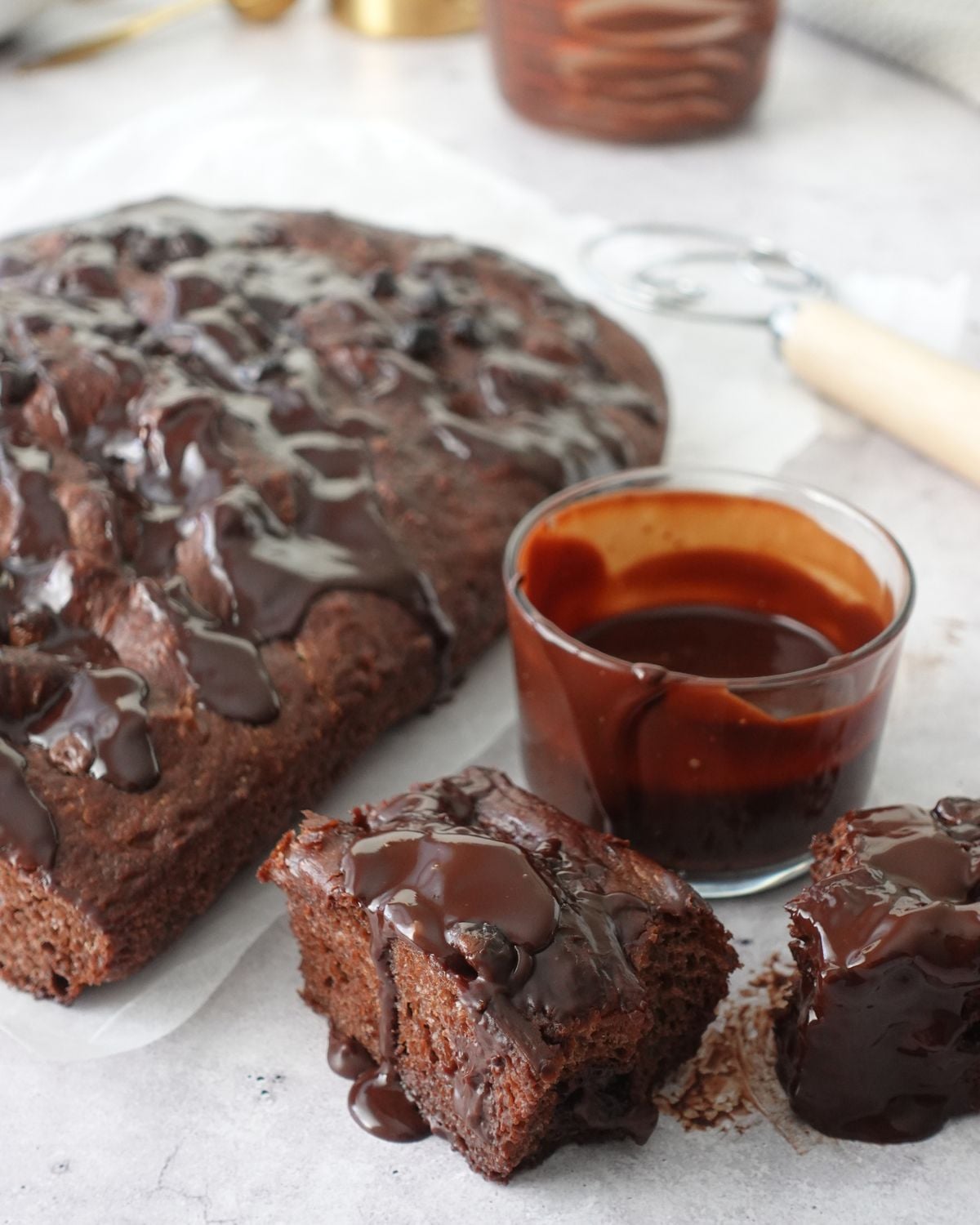 Same-day chocolate focaccia on parchment paper with ganache drizzled over the top, two slices cut showing the chocolate crumb, and a glass of ganache on the side