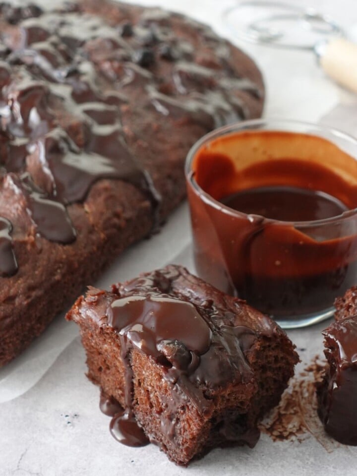 Same-day chocolate focaccia on parchment paper with ganache drizzled over the top, two slices cut showing the chocolate crumb, and a glass of ganache on the side