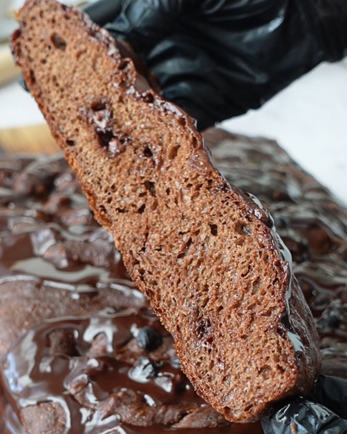 A gloved hand lifting a slice of chocolate focaccia showing the thick airy chocolate crumb with melted chocolate chips, with ganache pooling over the top on a wooden board