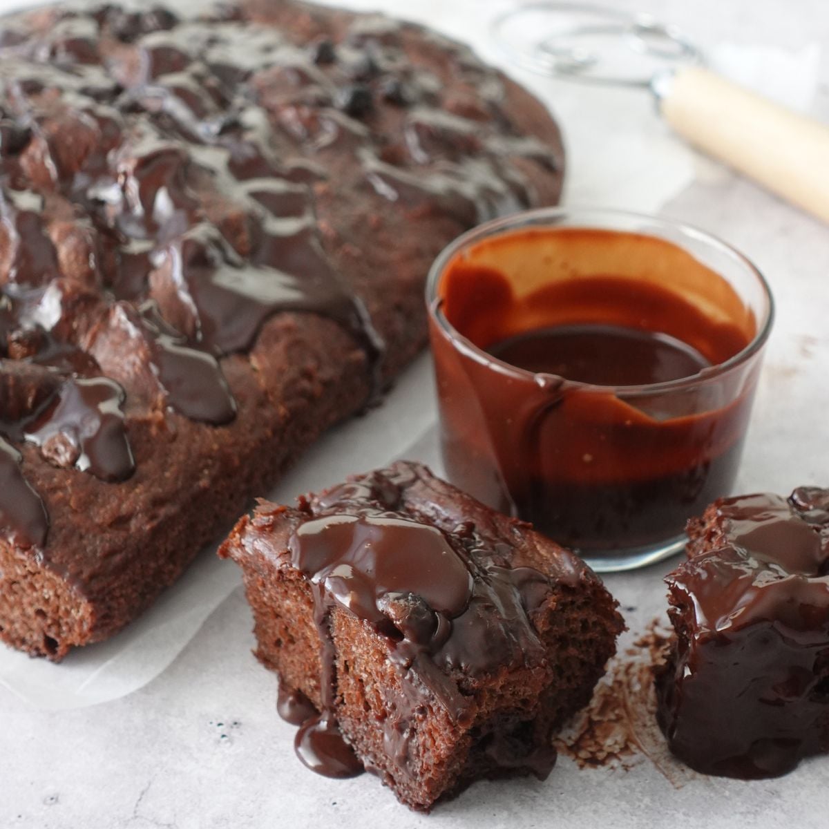 Same-day chocolate focaccia on parchment paper with ganache drizzled over the top, two slices cut showing the chocolate crumb, and a glass of ganache on the side