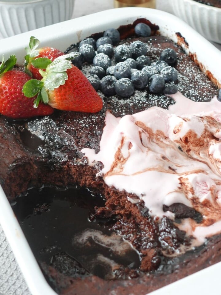 Chocolate self saucing pudding topped with strawberries, blueberries and ice cream, with a spoon through the center showing the sauce underneath