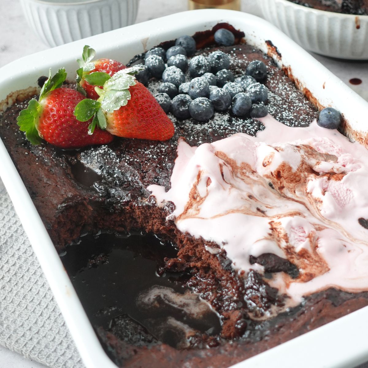 Chocolate self saucing pudding topped with strawberries, blueberries and ice cream, with a spoon through the center showing the sauce underneath
