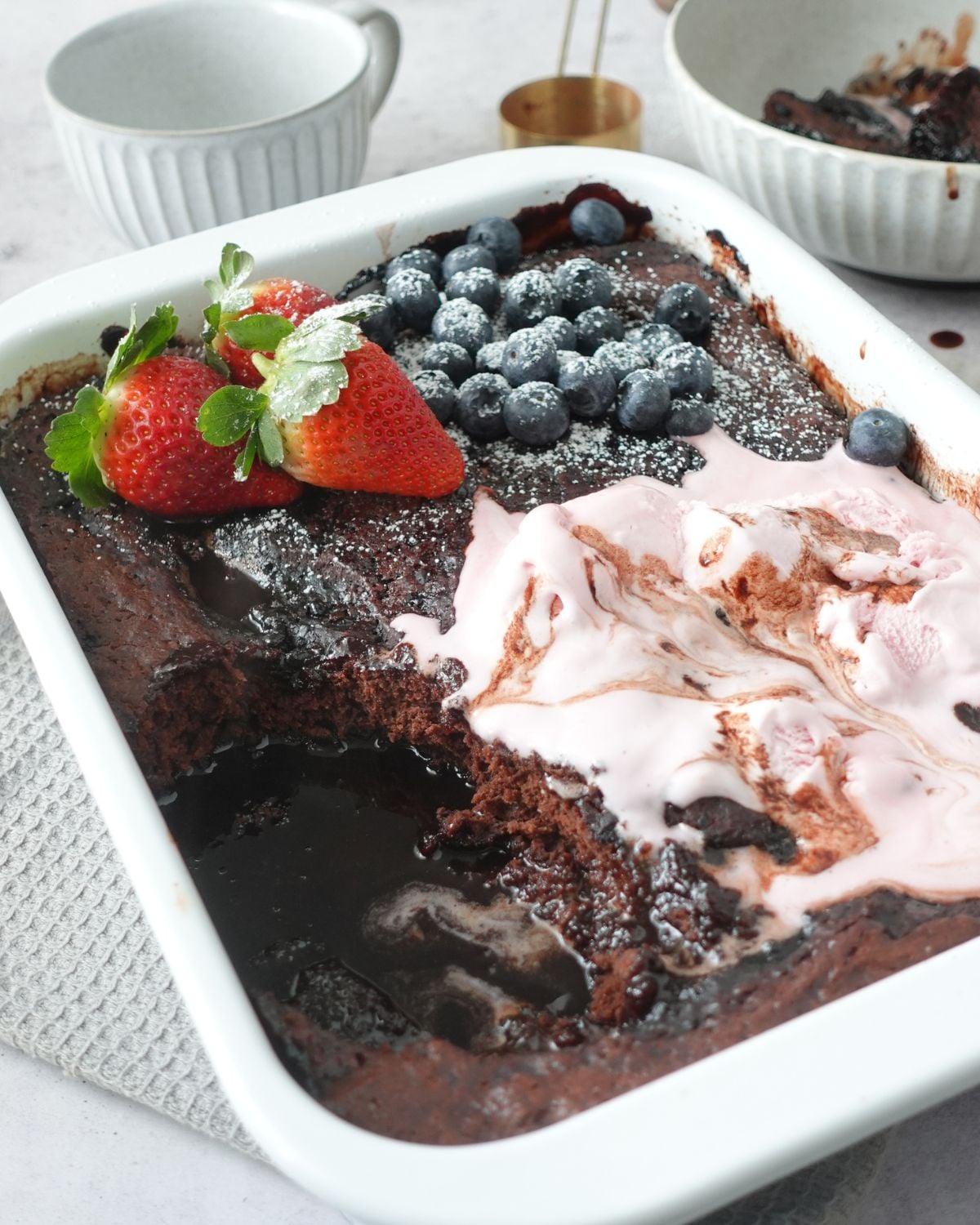 Chocolate self saucing pudding topped with strawberries, blueberries and ice cream, with a spoon through the center showing the sauce underneath