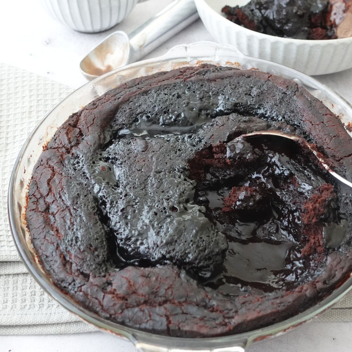 Chocolate self saucing pudding in a pie dish with a spoon through the center showing the sauce underneath