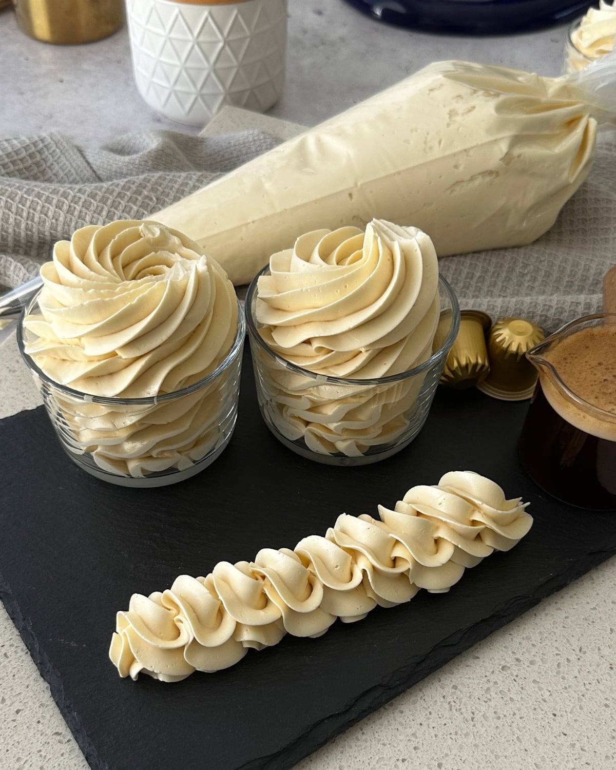Coffee hot water buttercream piped into swirls in glass jars with a rope border and espresso pods on a slate board