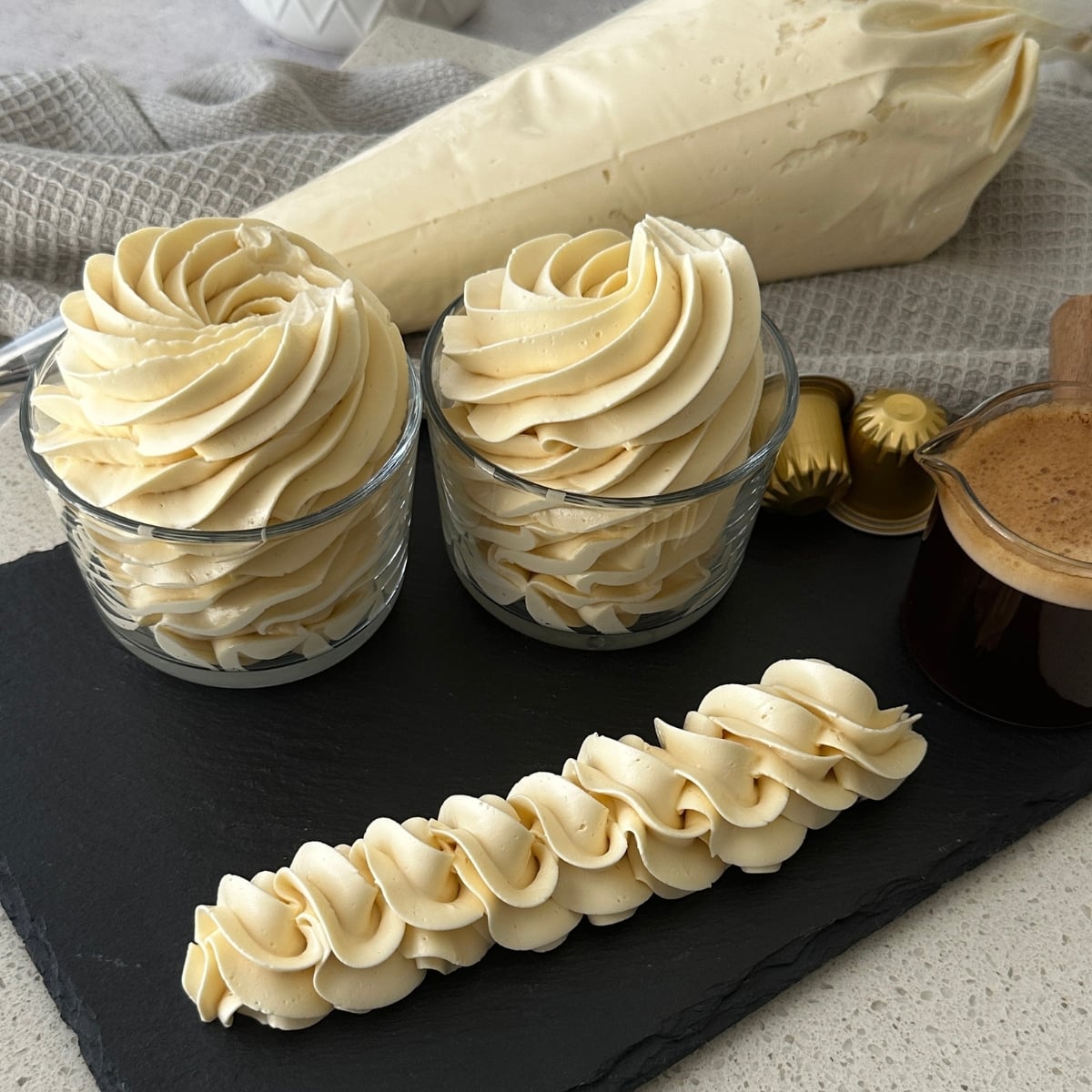 Coffee hot water buttercream piped into swirls in glass jars with a rope border and espresso pods on a slate board