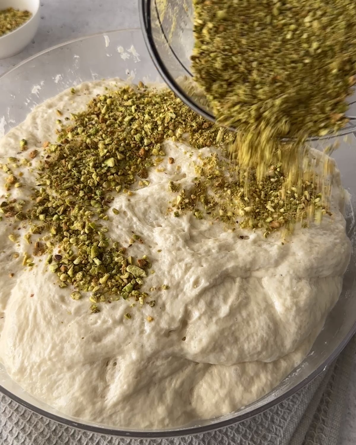 Crushed pistachios being poured over risen focaccia dough in a glass bowl