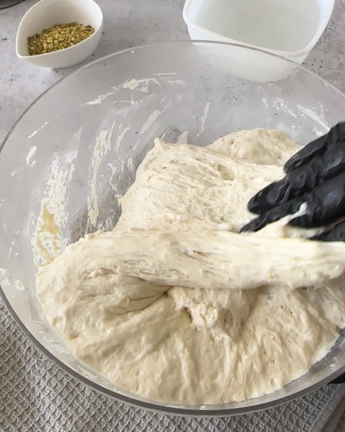 Gloved hand stretching and folding focaccia dough in a glass bowl