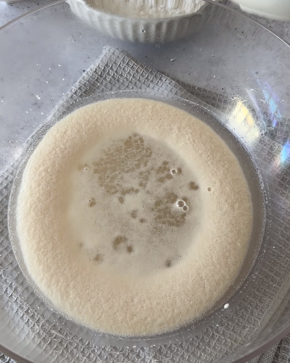 Foamy yeast mixture in a glass bowl with olive oil being poured in and a bowl of bread flour behind