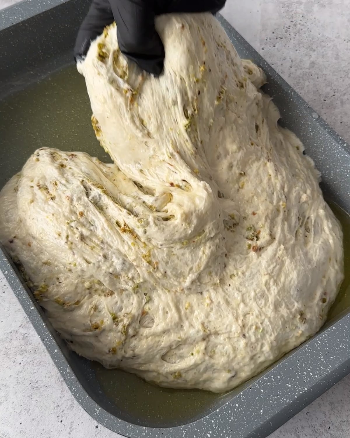 Gloved hand folding pistachio-studded dough in an oiled baking pan