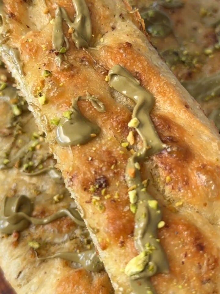 Slice of pistachio focaccia resting on the rest of the bake, topped with drizzled pistachio cream and crushed pistachios