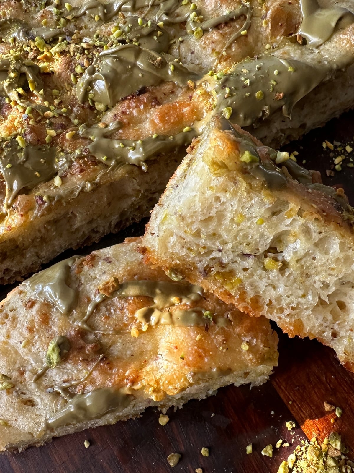 Pistachio focaccia slices on a wooden board showing the airy crumb with crushed pistachios through the dough and pistachio cream drizzled on top