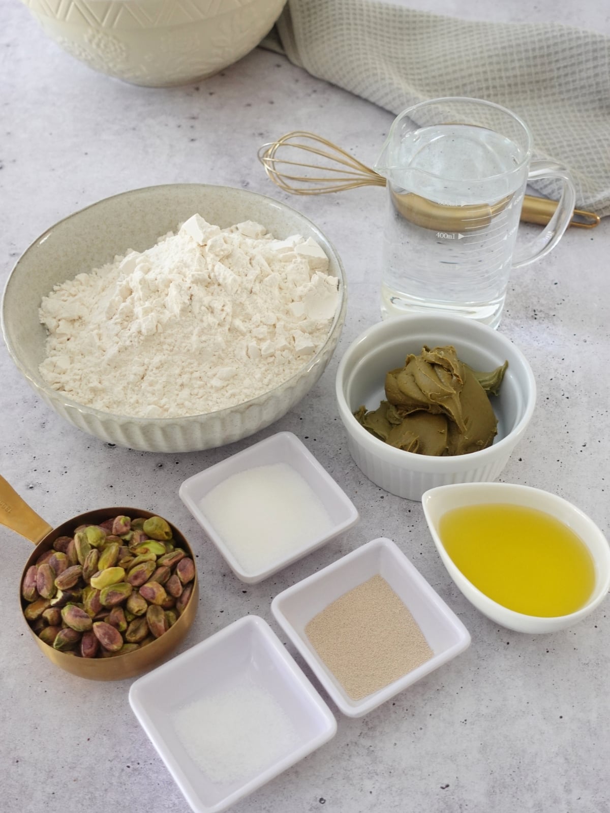 Ingredients for sweet pistachio focaccia including bread flour, warm water, shelled pistachios, pistachio cream, sugar, salt, instant dry yeast, and olive oil on a grey surface