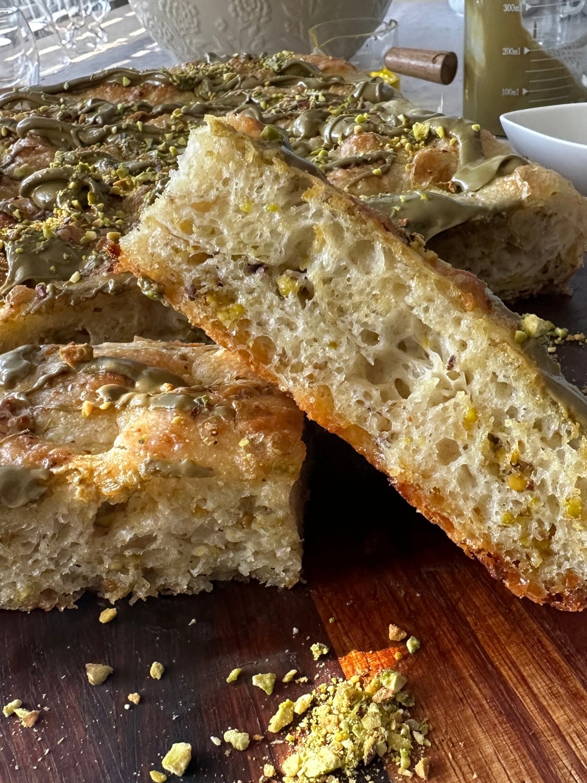 Thick slice of sweet focaccia propped on a wooden board, showing an airy crumb studded with crushed pistachios, with pistachio cream drizzled on top