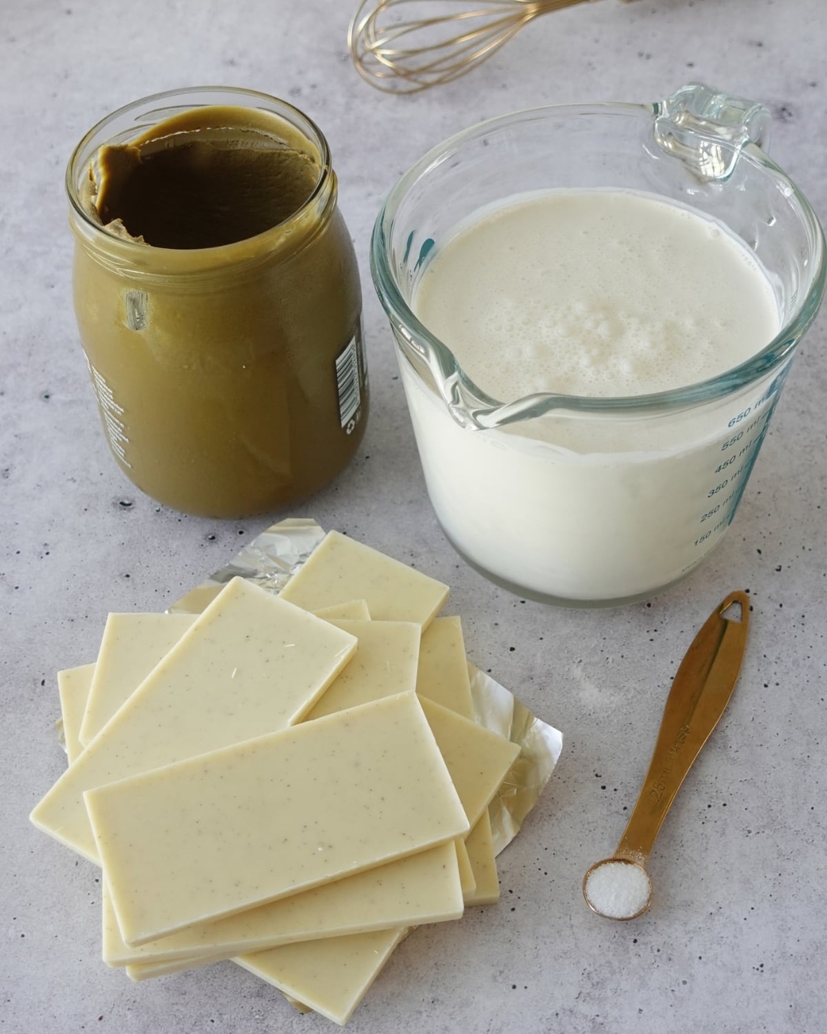 White chocolate bars, a jar of pistachio cream, a jug of cream and a measuring spoon of salt on a gray countertop