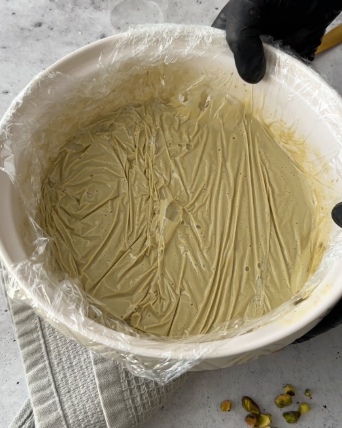 Gloved hands pressing cling wrap onto the surface of pistachio ganache in a white bowl