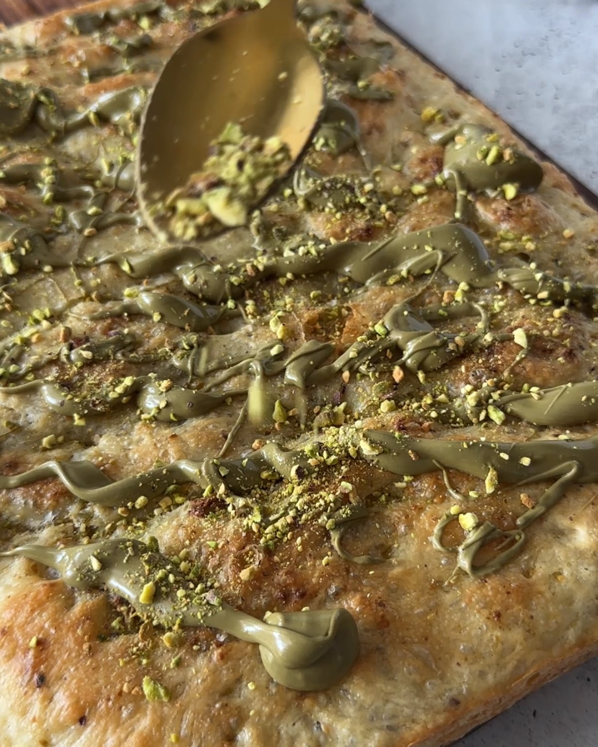 Spoon scattering crushed pistachios over baked focaccia drizzled with pistachio cream